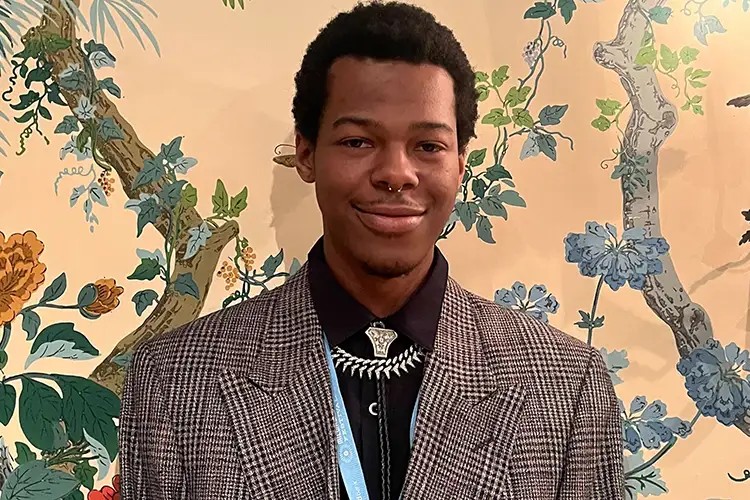 A person in a gray plaid blazer and black shirt stands in front of a floral wallpaper backdrop, smiling softly.
