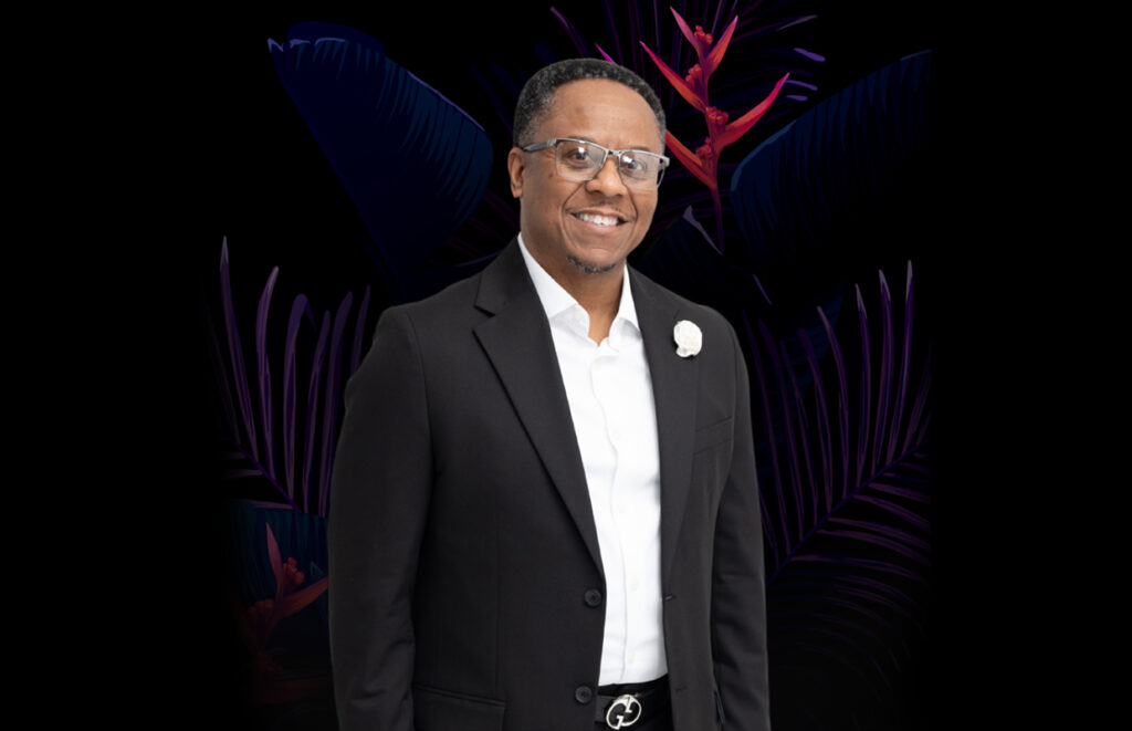 Smiling man in a black suit and white shirt posing against a dark tropical background with purple leaves.