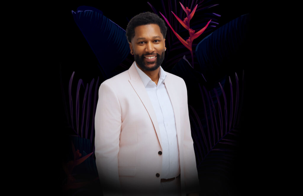 Smiling man in a light pink blazer and white shirt posing in front of dark tropical plants.