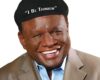 Smiling Black man in a tan blazer and light blue shirt wearing a black cap that reads "I Be Thinkin'"