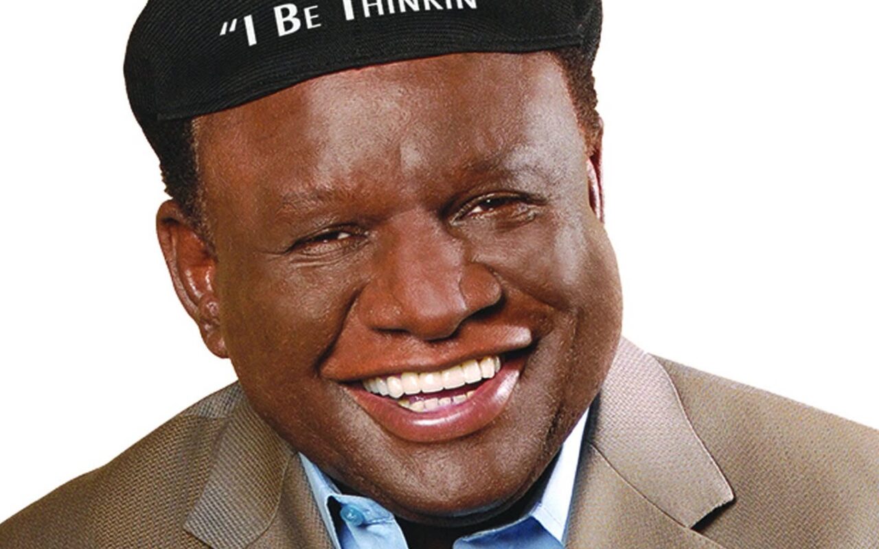 Smiling Black man in a tan blazer and light blue shirt wearing a black cap that reads "I Be Thinkin'"
